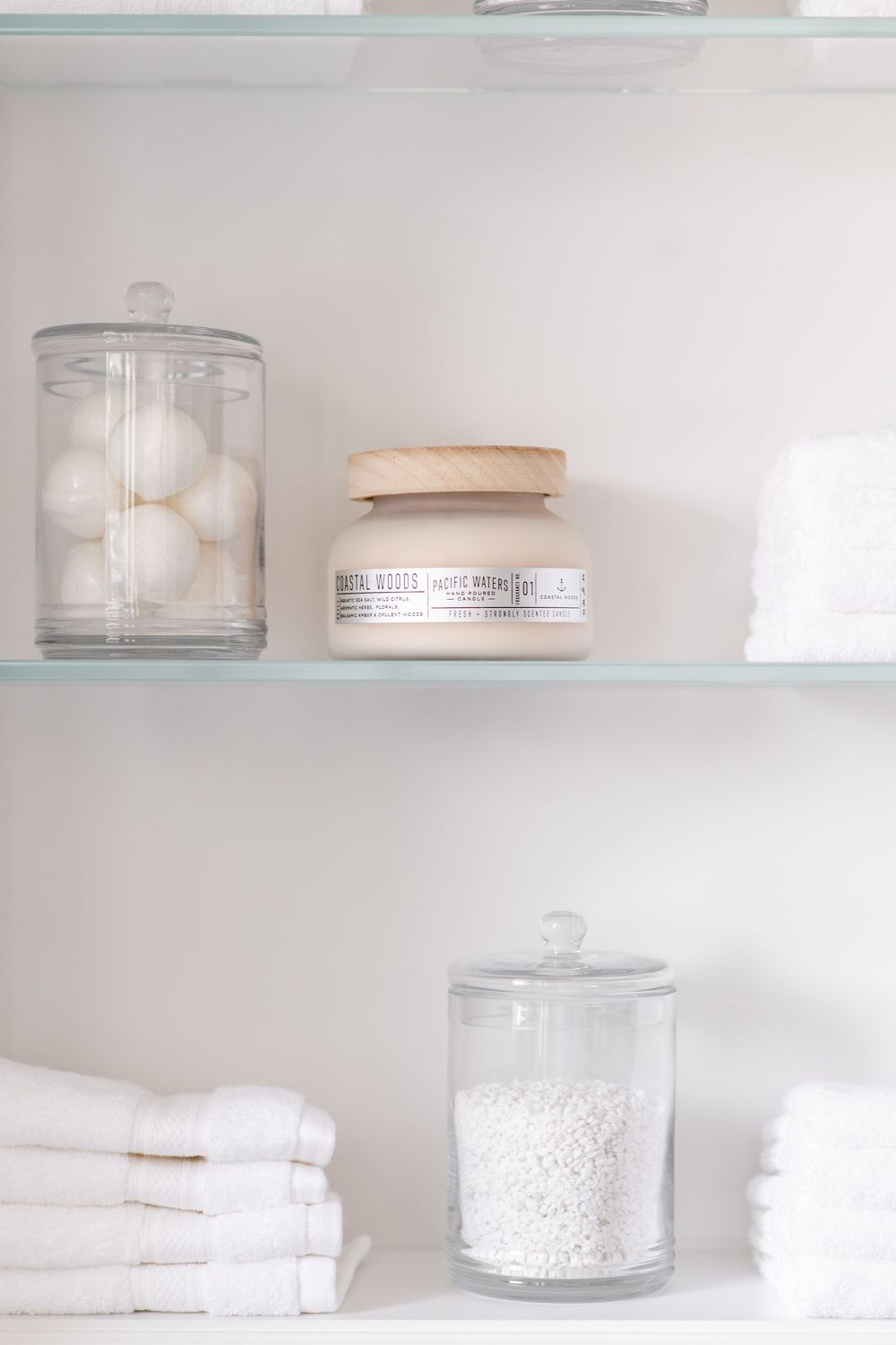 White bathroom shelf with glass jars holding cotton balls, folded white towels, and a cream candle jar.