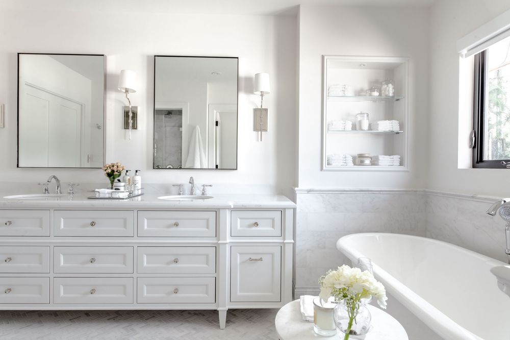 Elegant white bathroom with double vanity, marble countertop, dual mirrors, wall sconces, built-in shelving, and freestanding soaking tub.