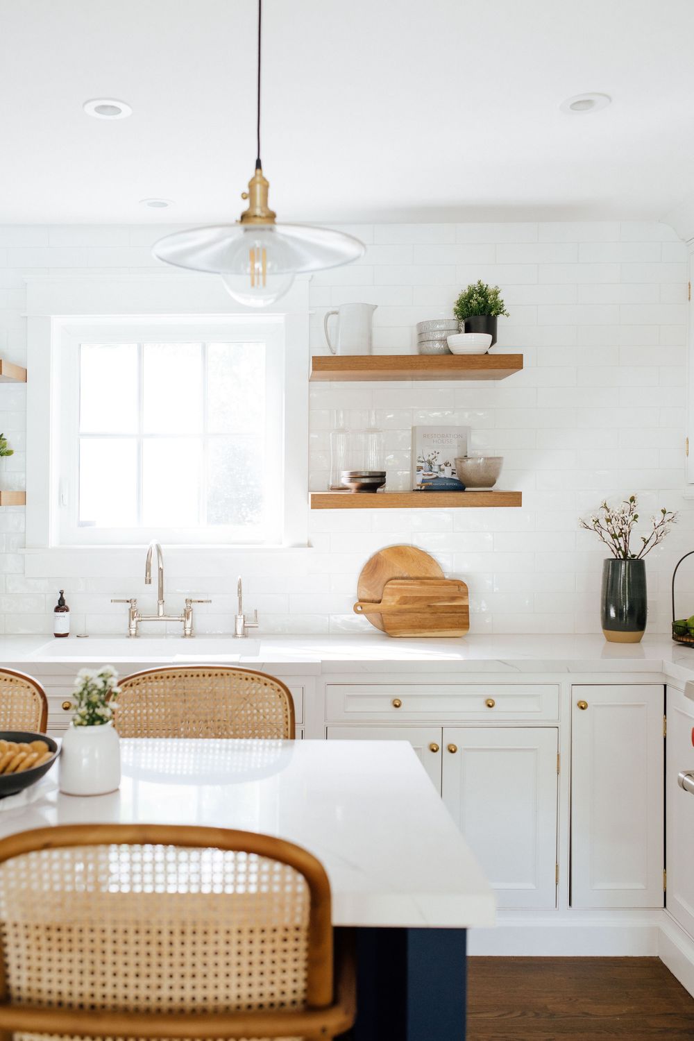 Bright white kitchen featuring open wood shelves, pendant light, farmhouse sink, and rattan bar stools with gold accents.