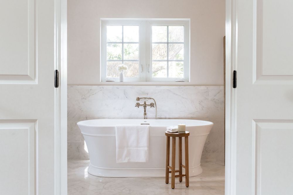 Elegant white freestanding bathtub in a serene bathroom with marble walls, wooden stool, and natural light from double windows.