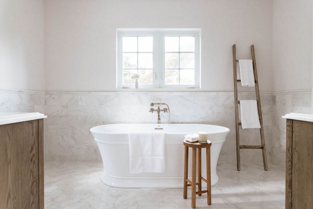 Elegant freestanding white bathtub in a marble-tiled bathroom with wooden vanities, a rustic ladder, and natural light.