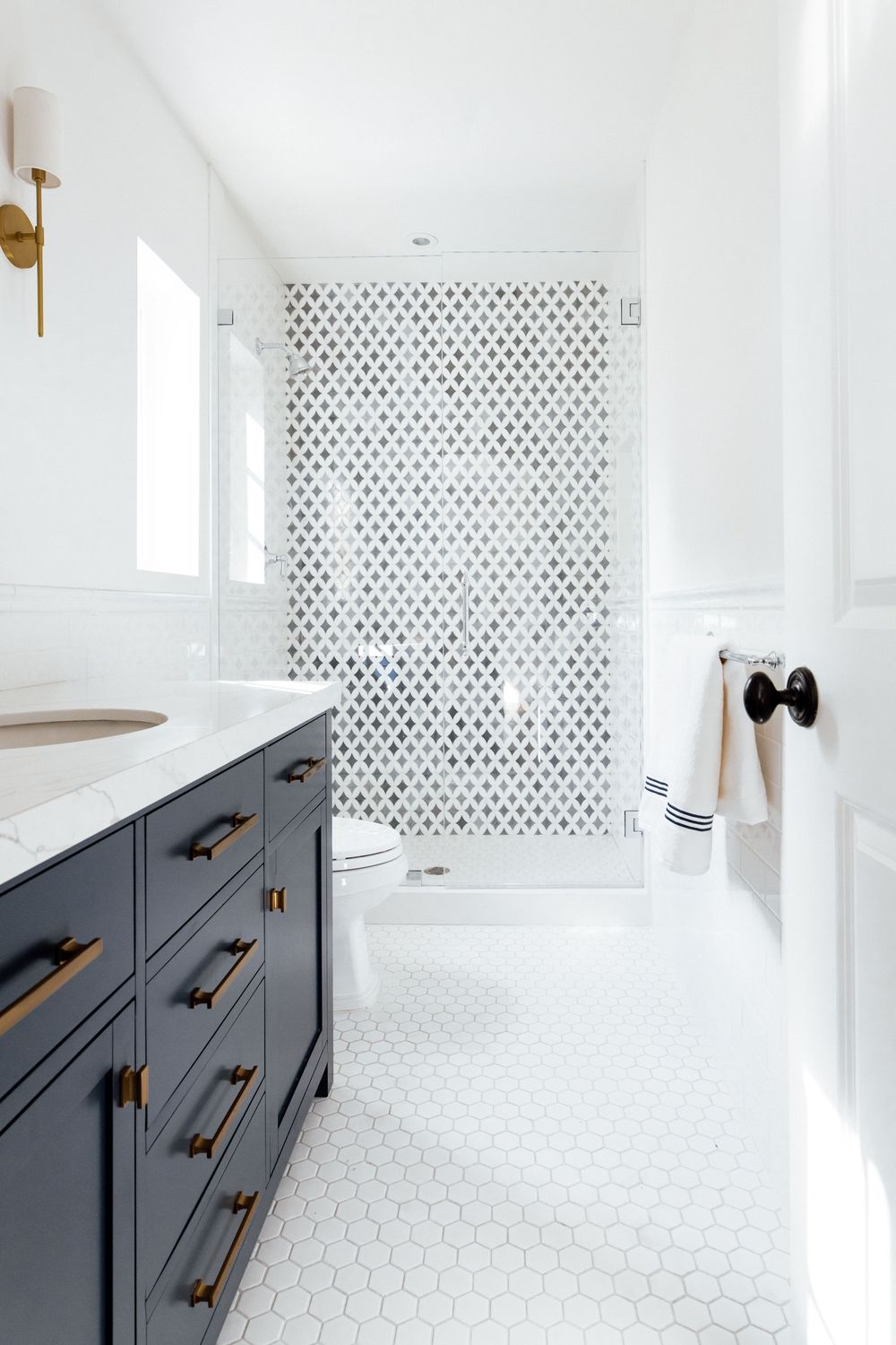 Navy blue bathroom vanity with brass pulls, white hex tile floor, and patterned black and white shower tile.