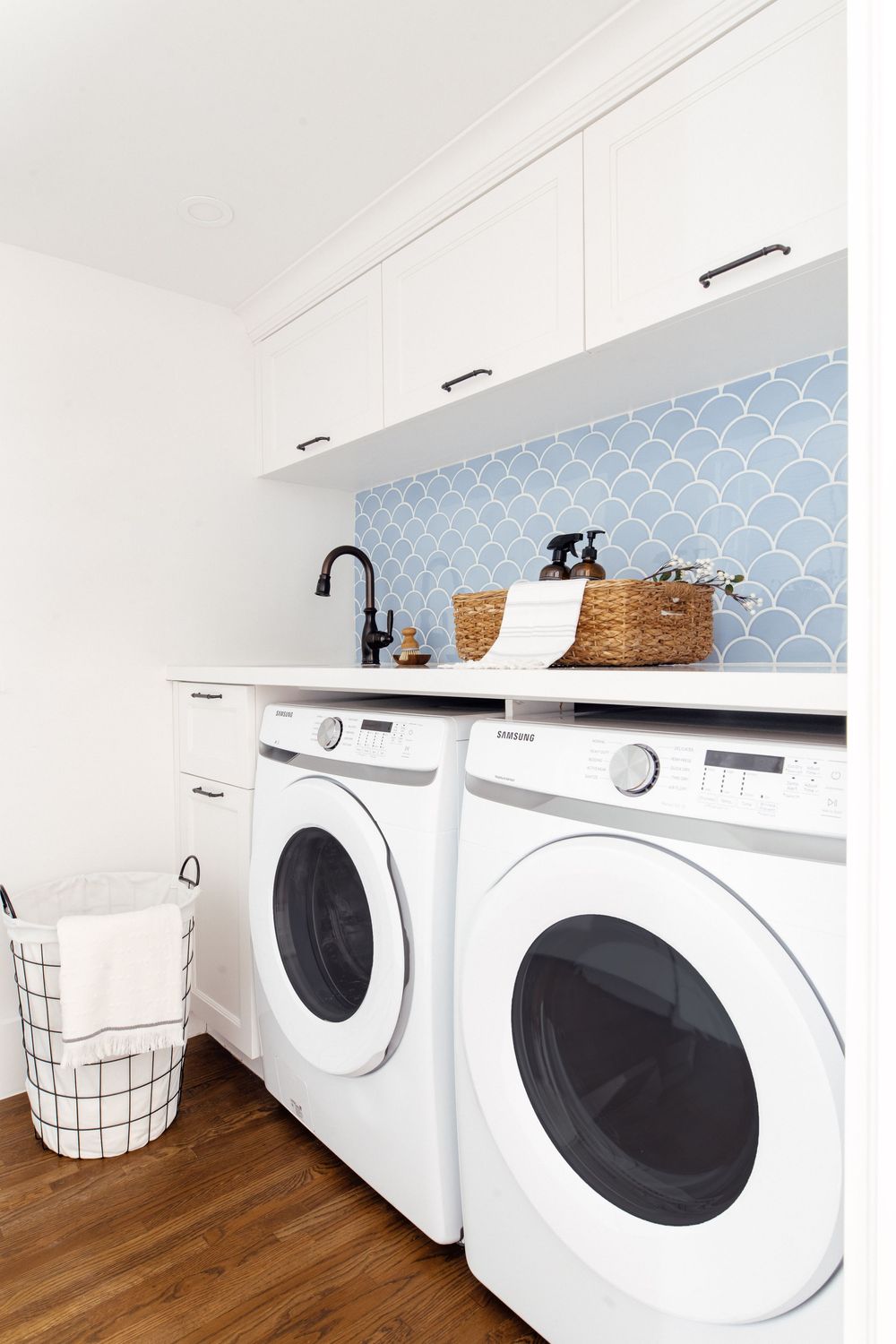 Modern white laundry room with front-load washer and dryer, blue fish scale tile backsplash, and upper cabinets.