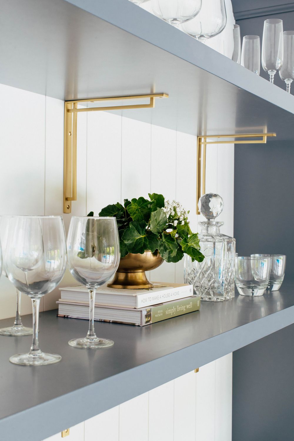 Gray shelves display wine glasses, a gold bowl with greenery, crystal decanter, and books with brass bracket supports.