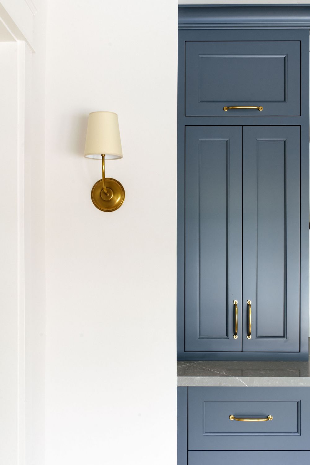 Blue-gray kitchen cabinets with gold brass hardware and marble countertop beside a brass wall sconce light fixture.