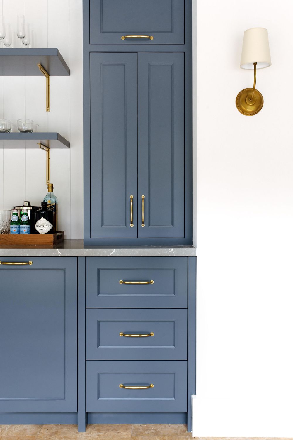 Blue shaker kitchen cabinets with brass hardware and marble countertop beside open shelving with gold wall sconce.