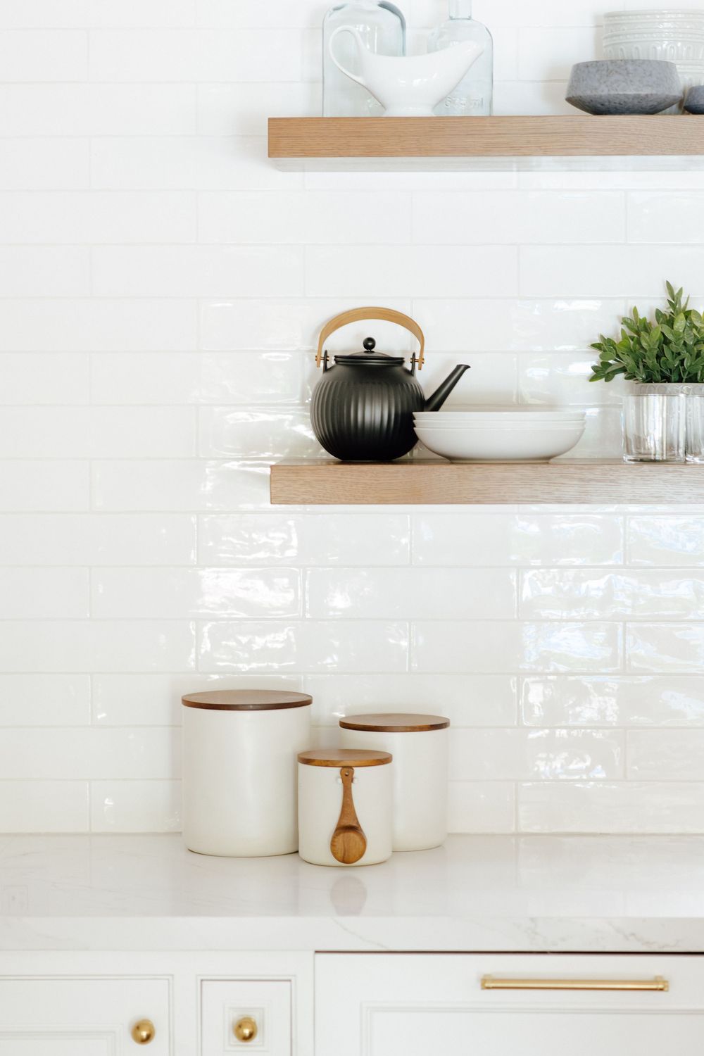 White kitchen shelves with wood brackets hold a black kettle, bowl, and plant above white canisters with wood lids.