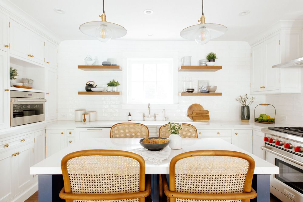 Bright white kitchen with cane dining chairs, open wood shelves, marble island, and gold pendant lights.