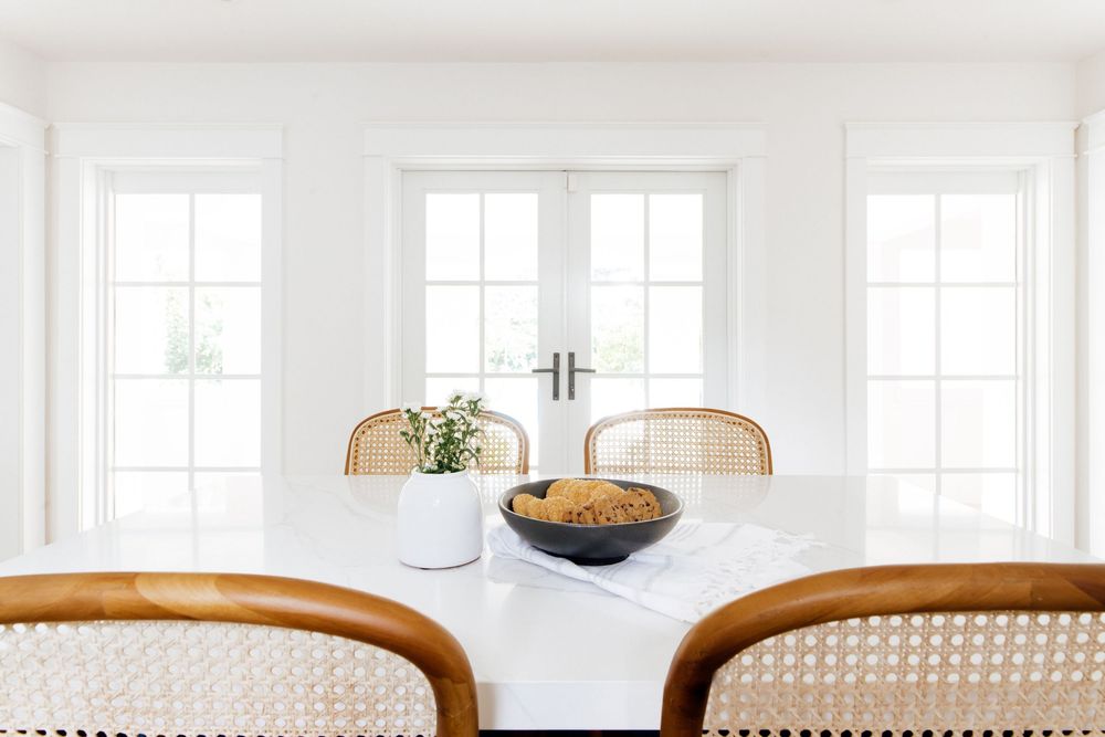 White dining room with marble table, rattan chairs, and French doors in a bright minimalist space.