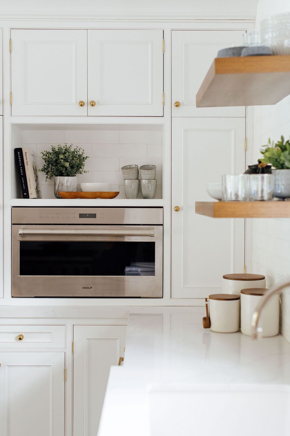 White kitchen with gold hardware, built-in Wolf oven, open shelving, and white shaker cabinets with subway tile backsplash.