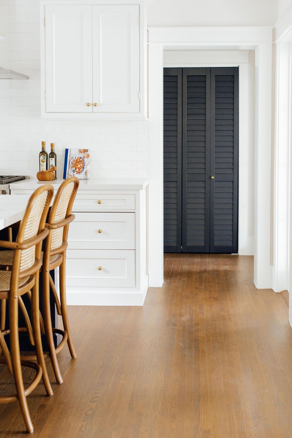 White kitchen with rattan bar stools, gold hardware drawers, and a dark navy blue louvered pantry door.