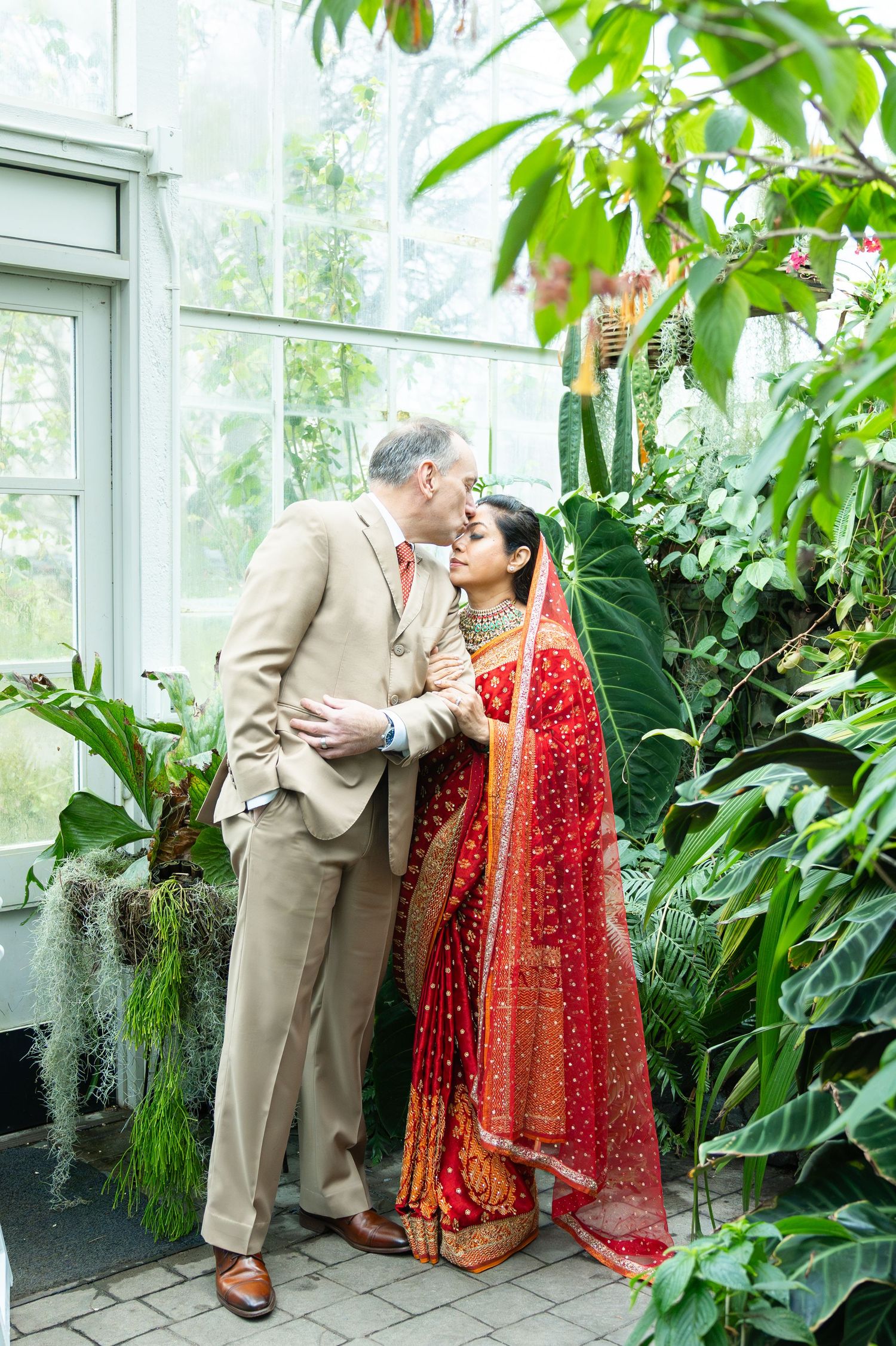 Couple in a greenhouse, groom in tan suit kissing bride wearing red Indian bridal saree surrounded by lush tropical plants.