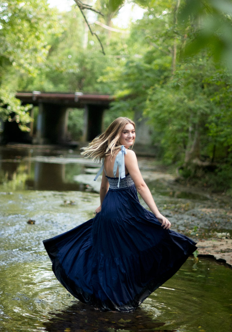 Young Black woman in white dress and cowboy boots leans on stone wall in lush garden setting.