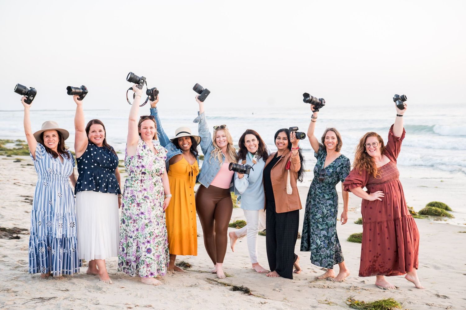Nine women photographers celebrating on a beach, raising cameras in the air, wearing colorful dresses with ocean backdrop.