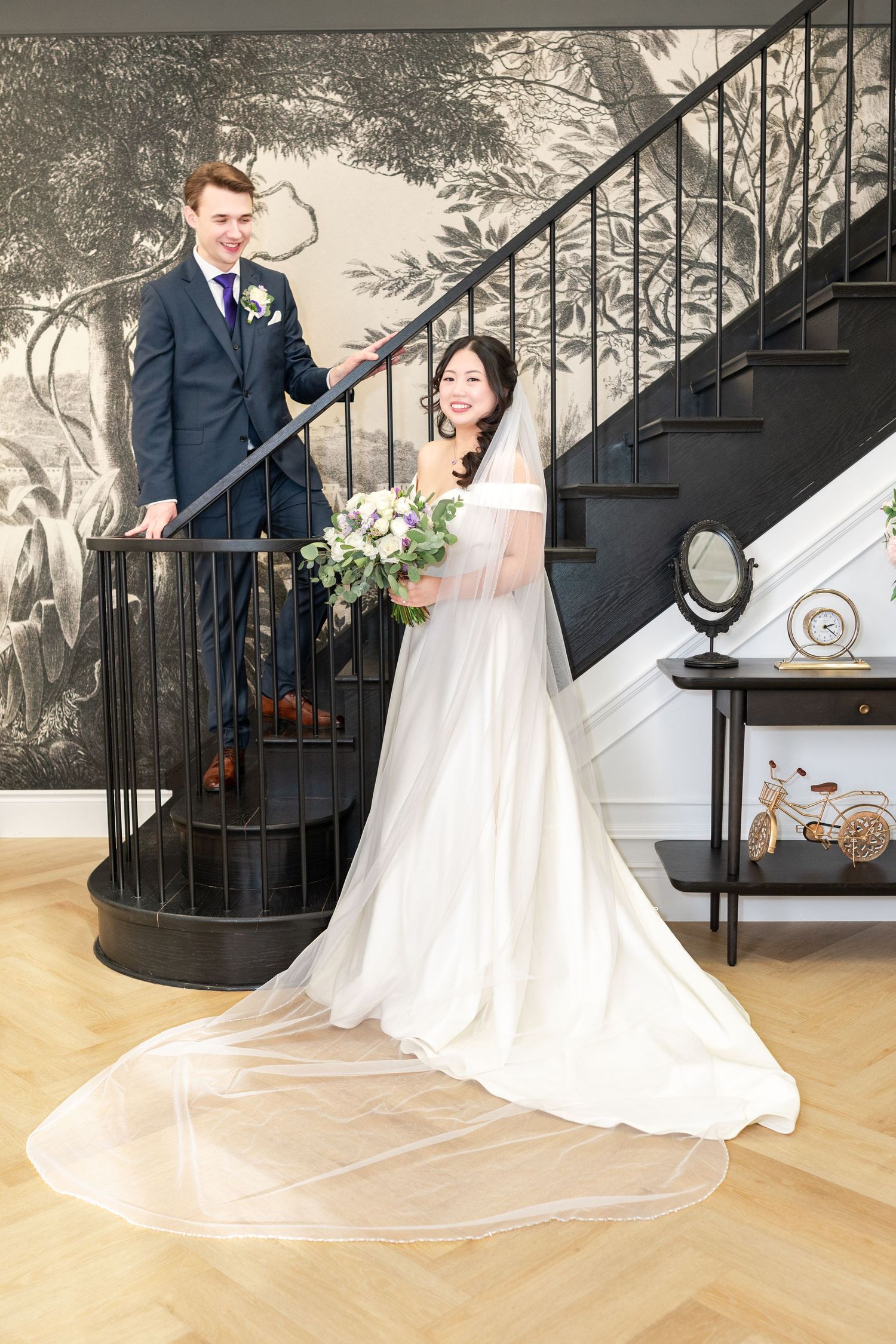 Elegant couple posing together, man in black tuxedo and woman in white hijab gown, holding hands in formal venue.