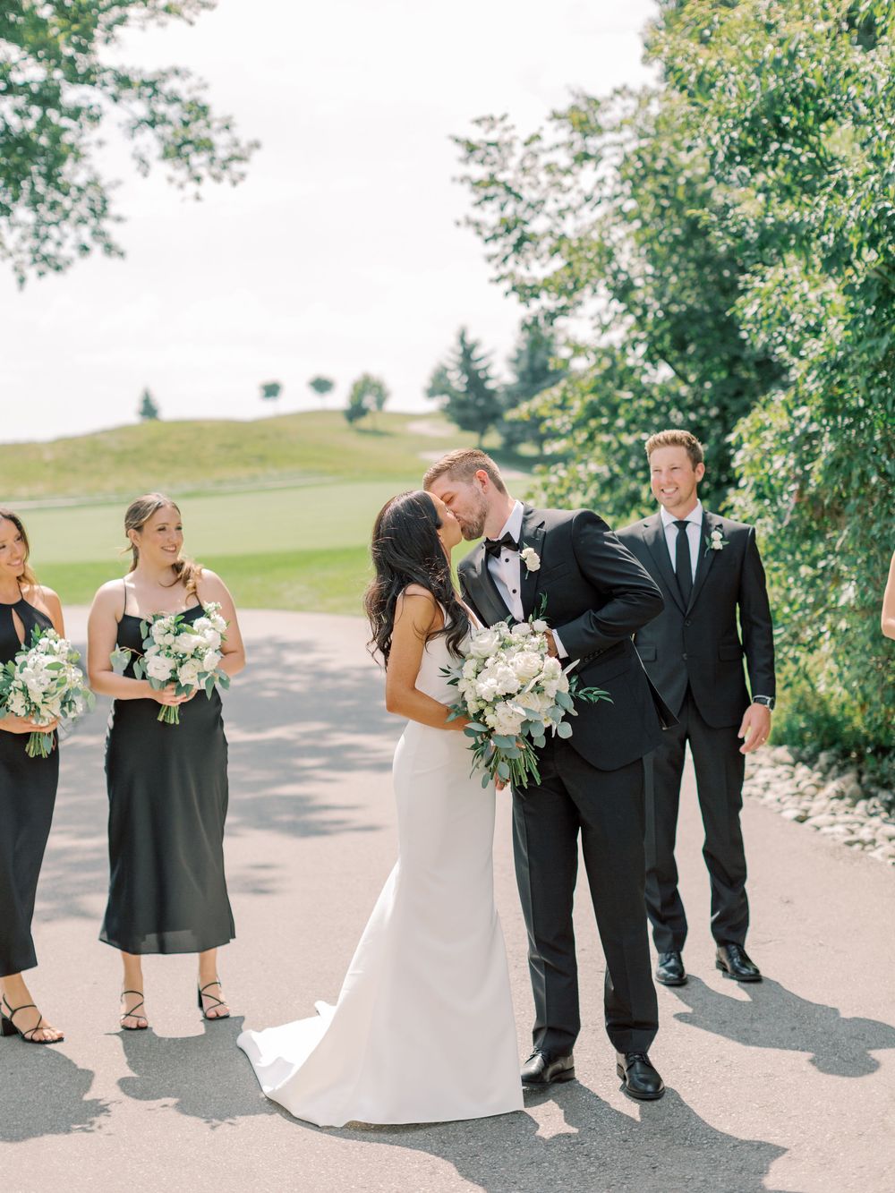 Bride and groom share a kiss surrounded by bridesmaids in black dresses and groomsmen at an outdoor wedding venue.
