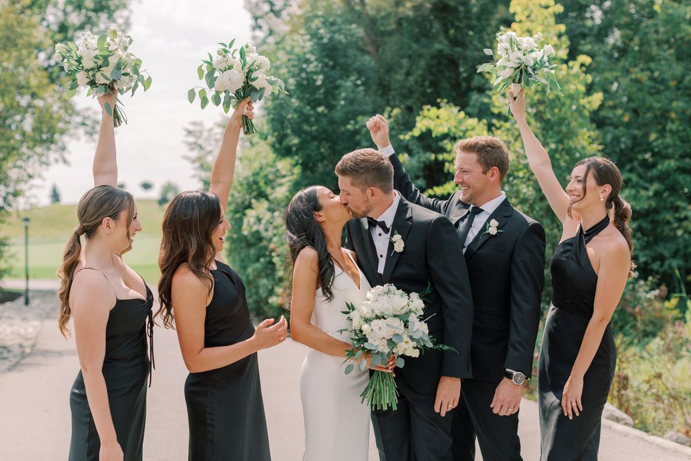 Wedding couple kissing surrounded by cheering bridesmaids in black dresses and groomsmen holding bouquets outdoors.