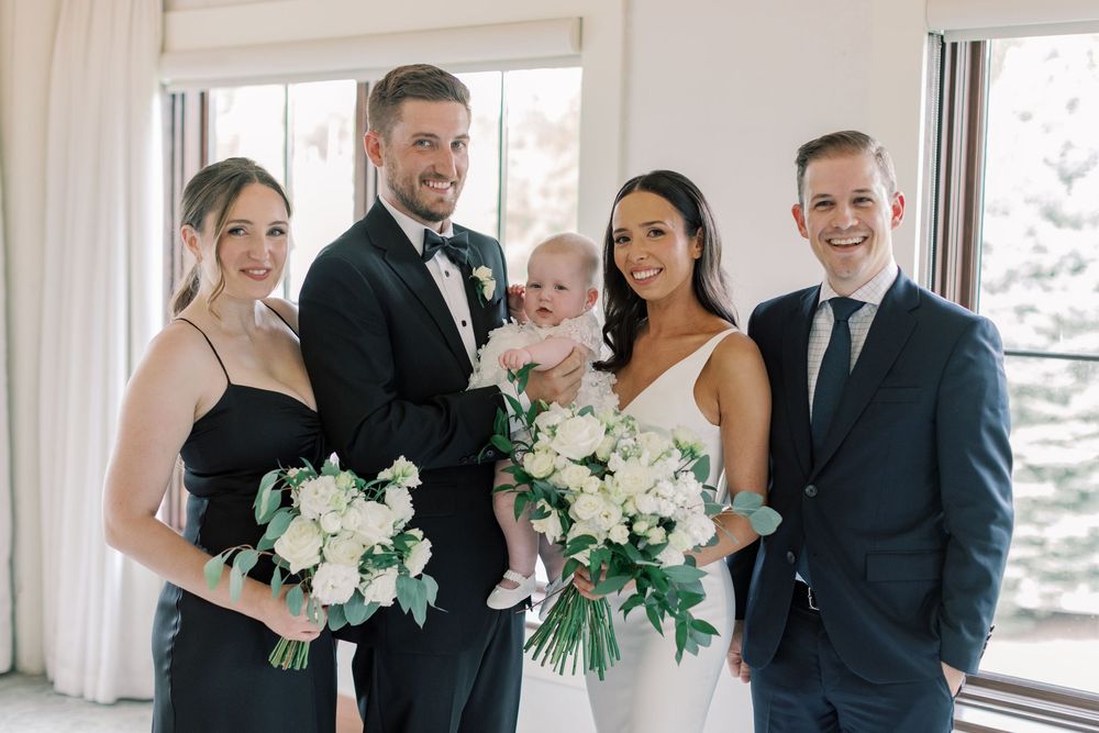 Wedding party poses indoors with bride holding bouquet, groom carrying baby, bridesmaid, and groomsman in formal attire.