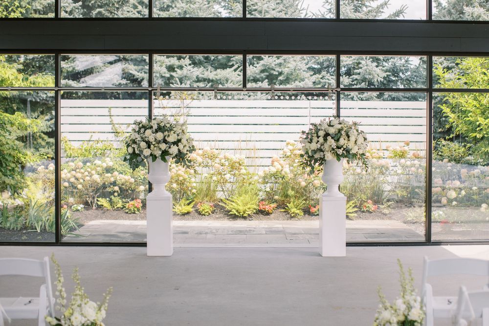 Elegant wedding ceremony setup with white floral arrangements on pedestals before large glass windows overlooking a lush garden.