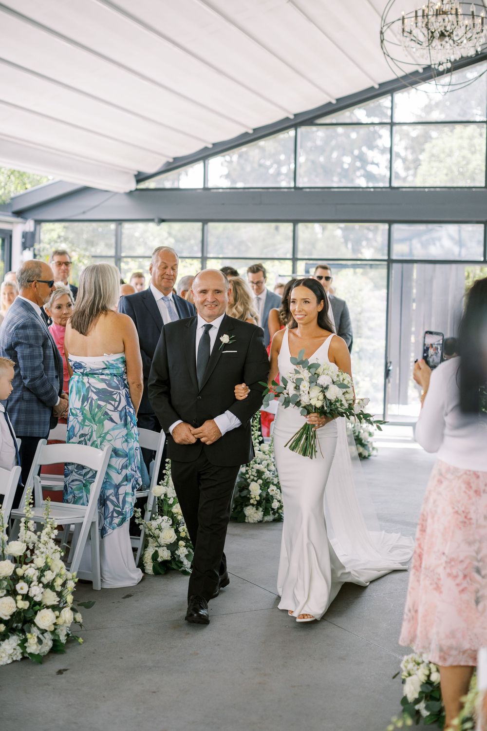Bride in white gown walks down aisle with escort at elegant indoor wedding ceremony with floral arrangements.
