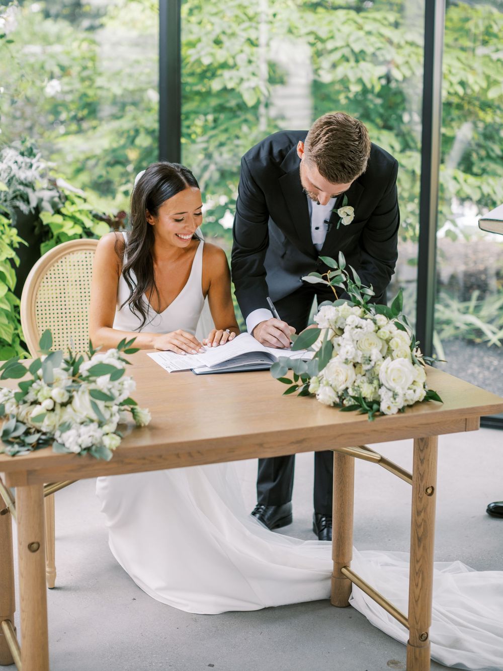 Bride and groom signing wedding registry at wooden table with white floral arrangements in elegant greenhouse venue.
