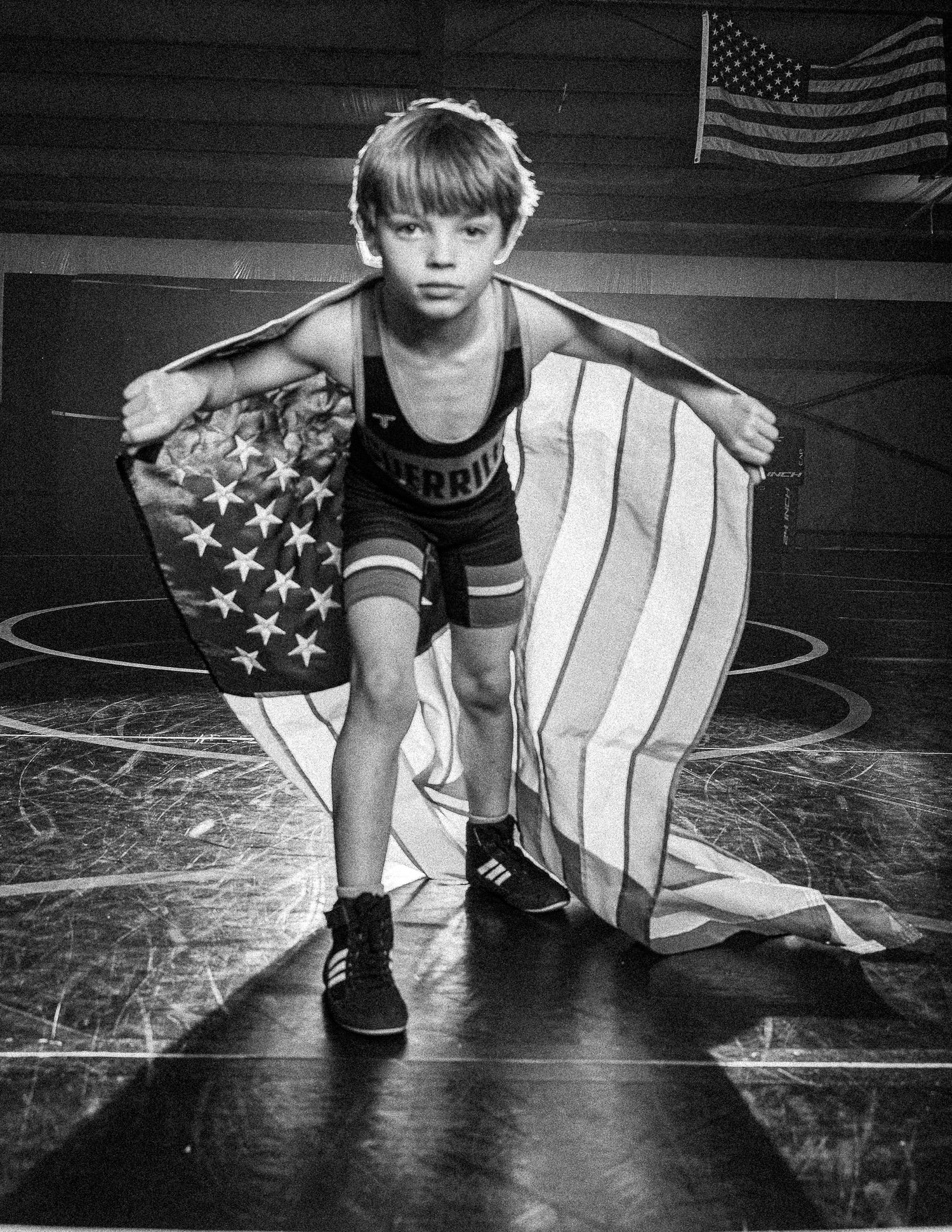 Young wrestler in singlet holds American flag on mat in black and white photo with US flag in background.
