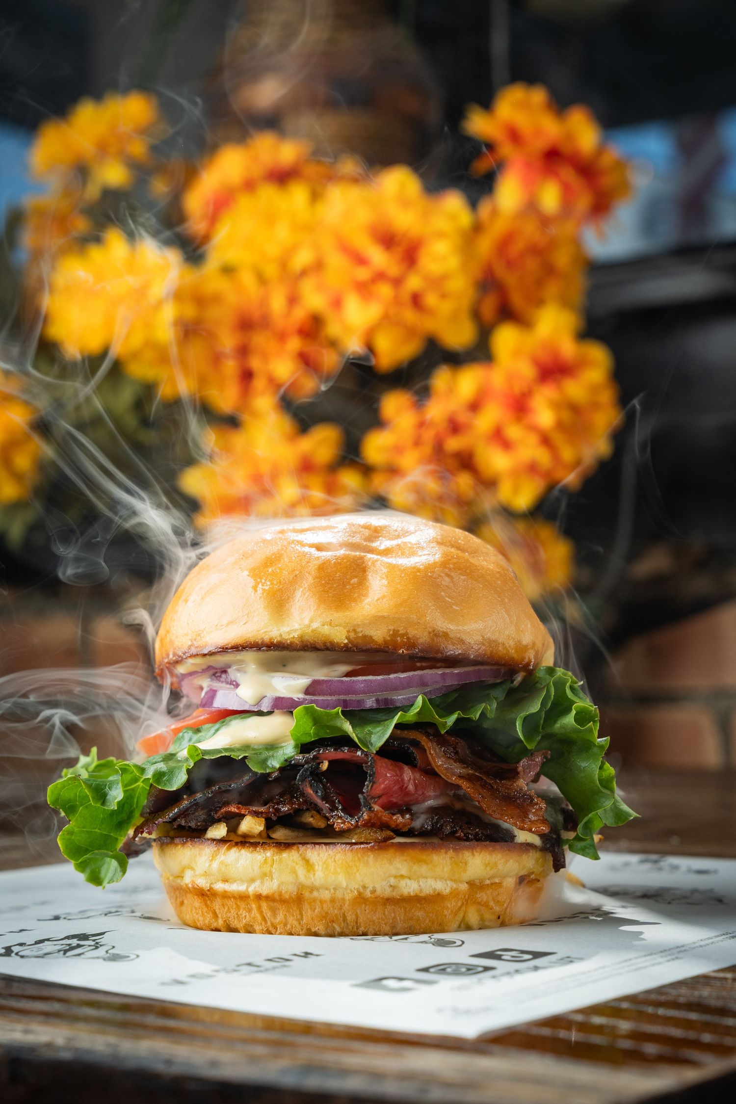 A steaming gourmet burger with lettuce, onion, and bacon on a brioche bun, set against vibrant yellow flowers.