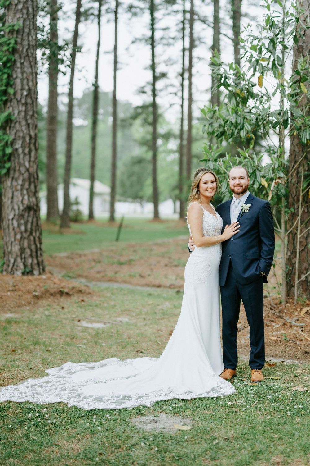 Bride and groom pose together in a pine forest, bride wearing a lace train wedding gown, groom in navy suit.