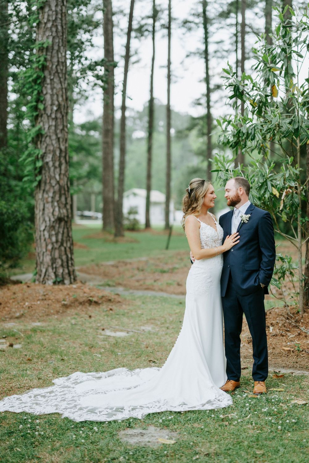 Bride in white gown with lace train stands with groom in navy suit outdoors among tall pine trees.