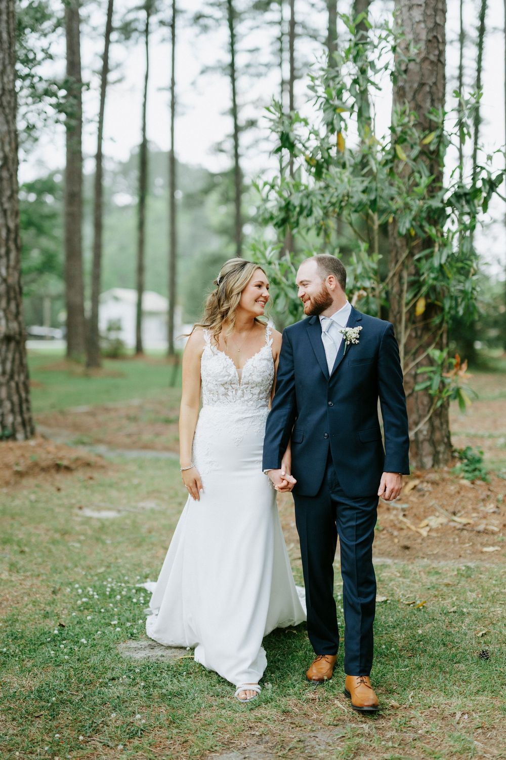 A bride and groom walk hand-in-hand through a pine tree forest, smiling at each other on their wedding day.