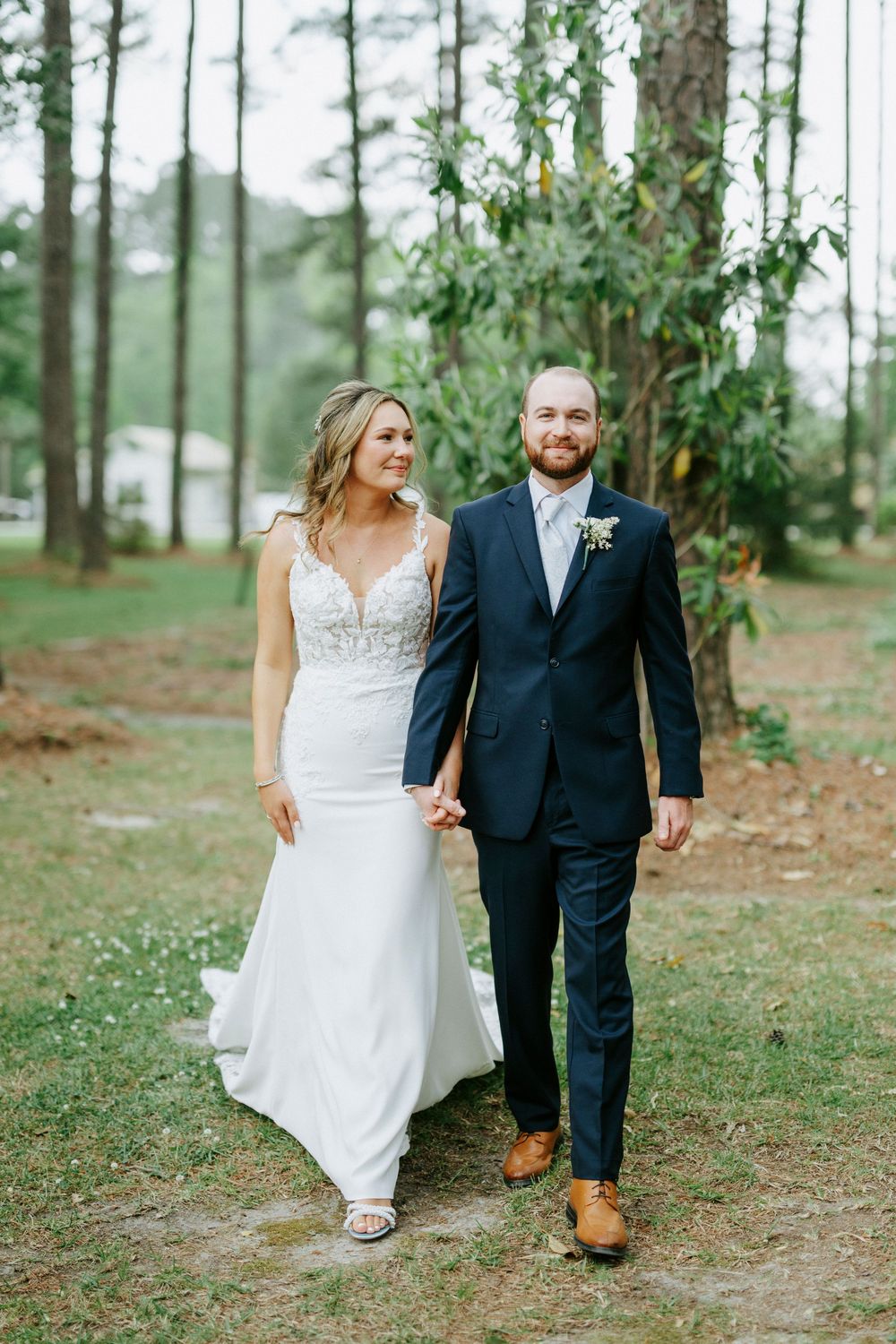 Bride in white gown and groom in navy suit hold hands walking outdoors among pine trees on their wedding day.