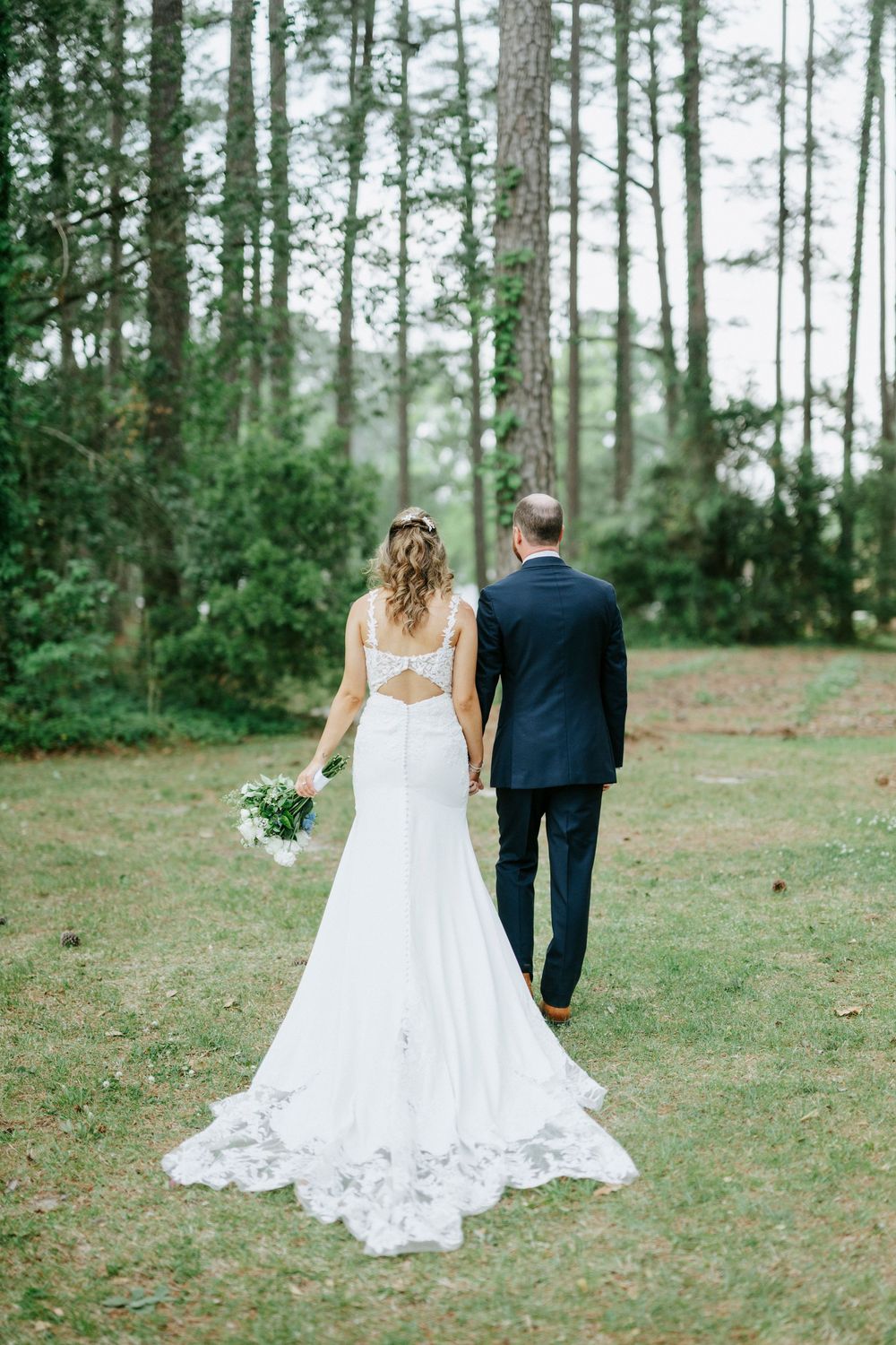 Bride and groom walk hand-in-hand through a pine forest, bride in backless lace wedding gown holding bouquet.