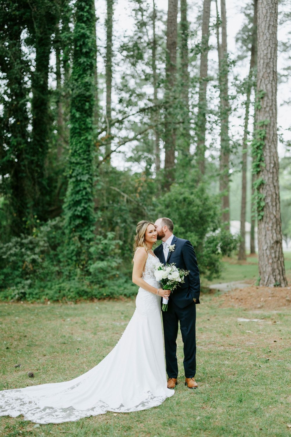 Bride and groom share a romantic moment in a lush forest setting, with the bride holding a white bouquet.