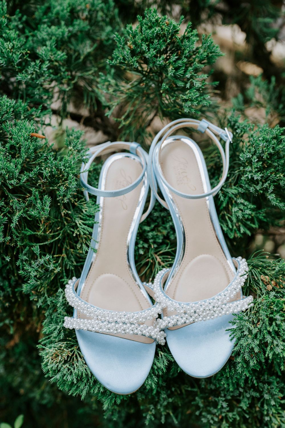 Light blue pearl-embellished wedding sandals with ankle straps resting on green evergreen branches.
