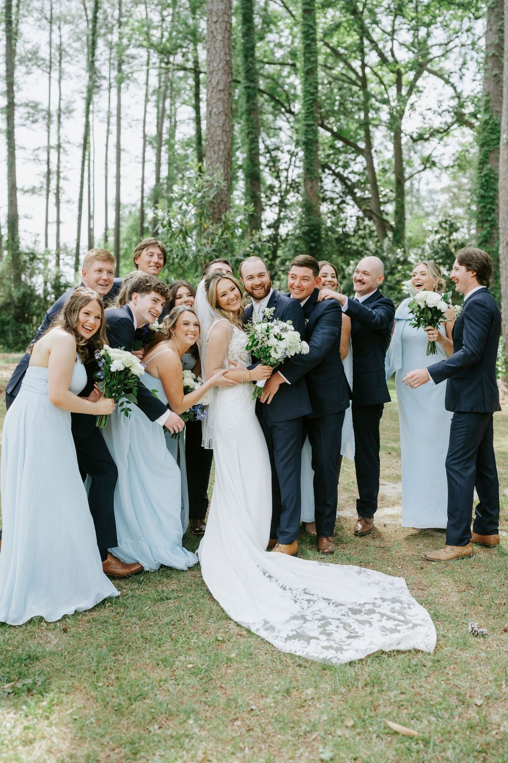 Wedding party posing together outdoors in a forest setting, bridesmaids in light blue gowns and groomsmen in navy suits.