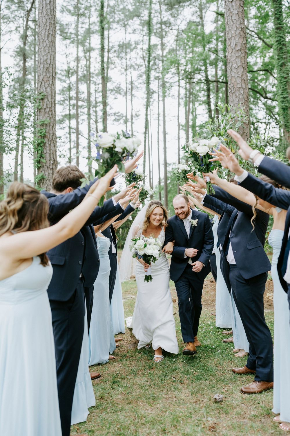 Bride and groom walk through arch of raised arms formed by wedding party in pine forest ceremony.