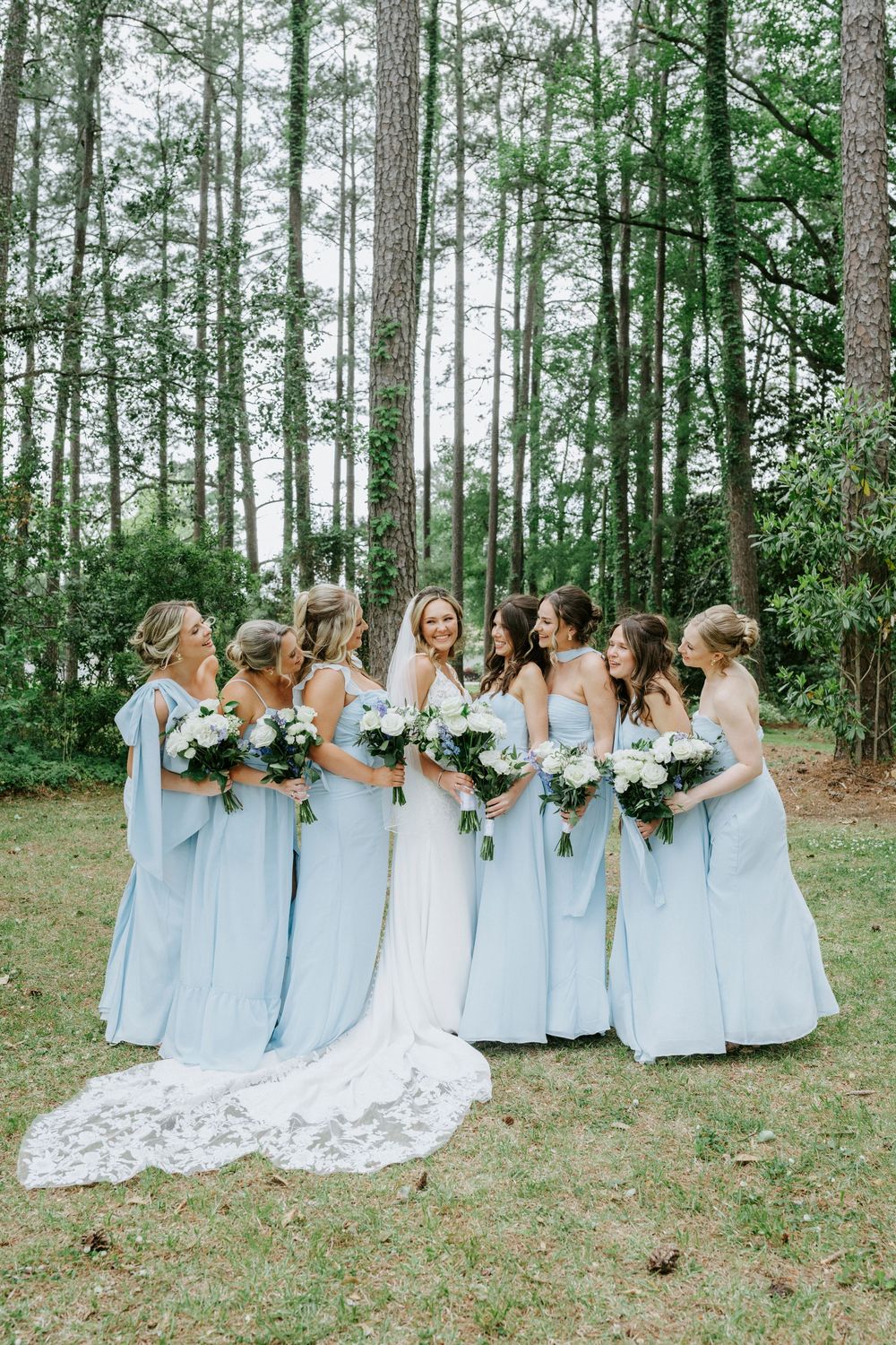 Bride in white gown with bridesmaids in light blue dresses holding white bouquets, posing outdoors among tall pine trees.
