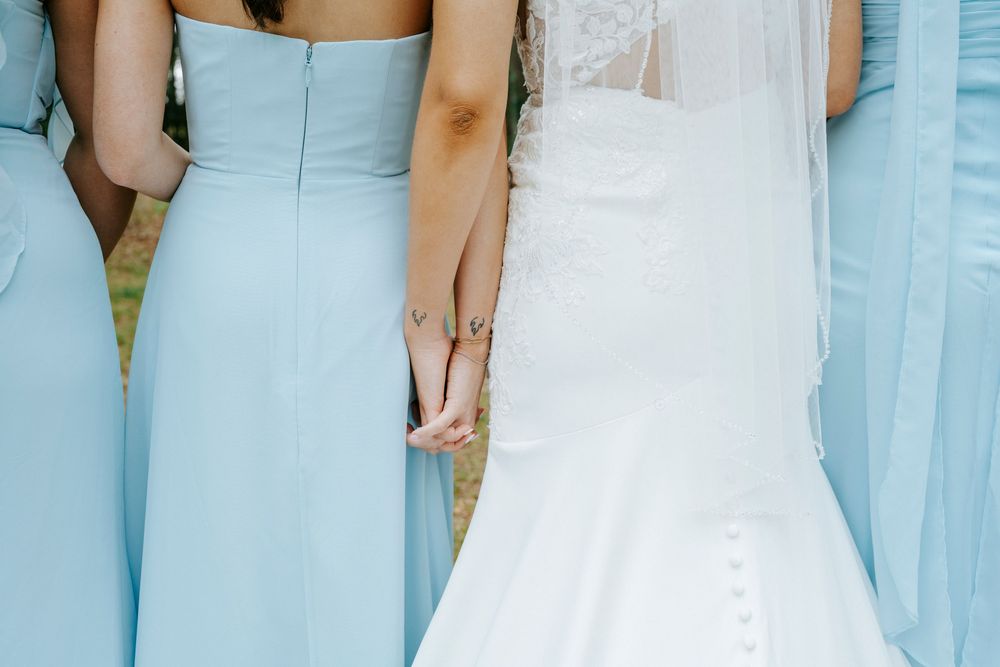 Bride in white lace gown stands with bridesmaids in light blue dresses, viewed from behind at an outdoor wedding.