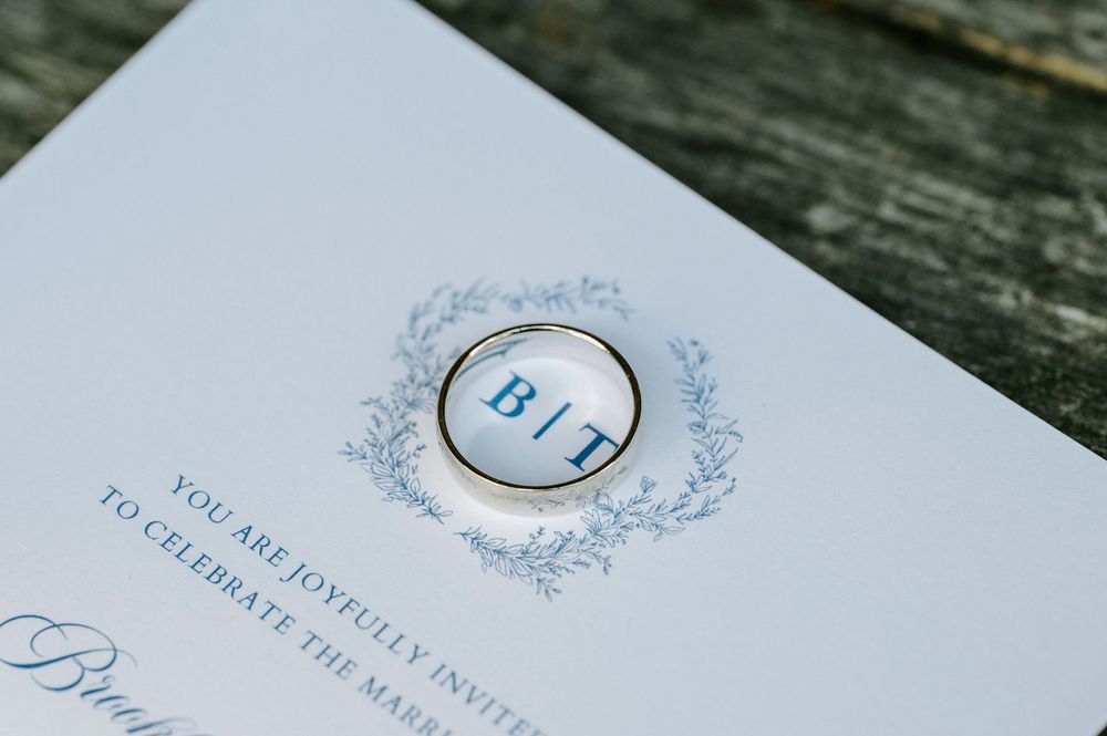 Wedding invitation with gold ring featuring monogram B and T initials crest design.
