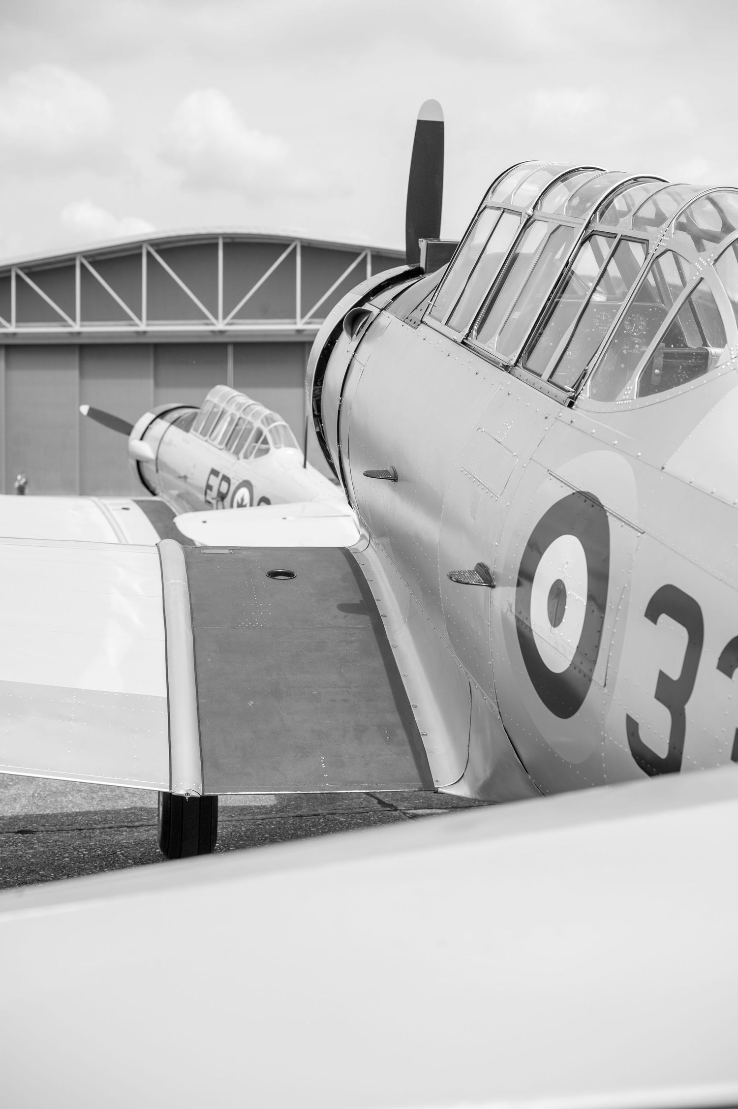 Black and white photo of a vintage WWII-era propeller aircraft with roundel markings parked on an airfield near a hangar.