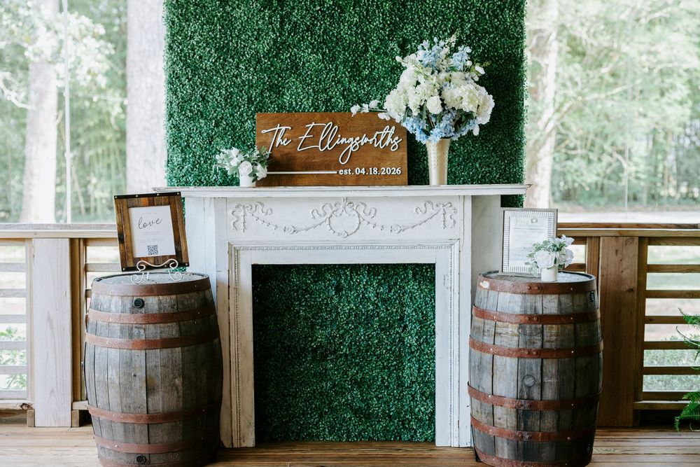 Wedding decor with white fireplace, greenery wall, wooden barrels, and floral arrangement at The Ellingtons reception.