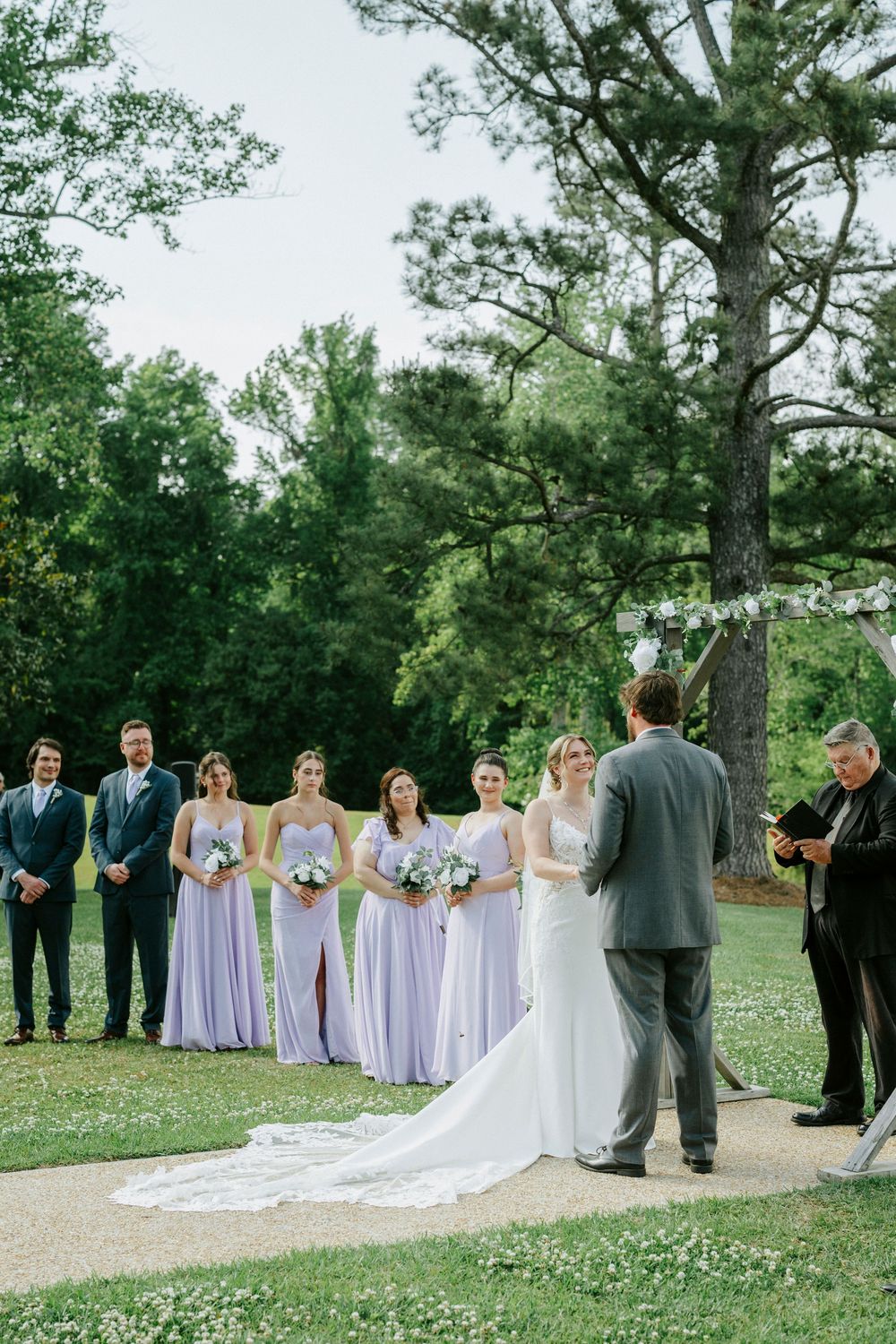 Outdoor wedding ceremony with bride in white gown, bridesmaids in lavender dresses, and groomsmen standing on lush green lawn.