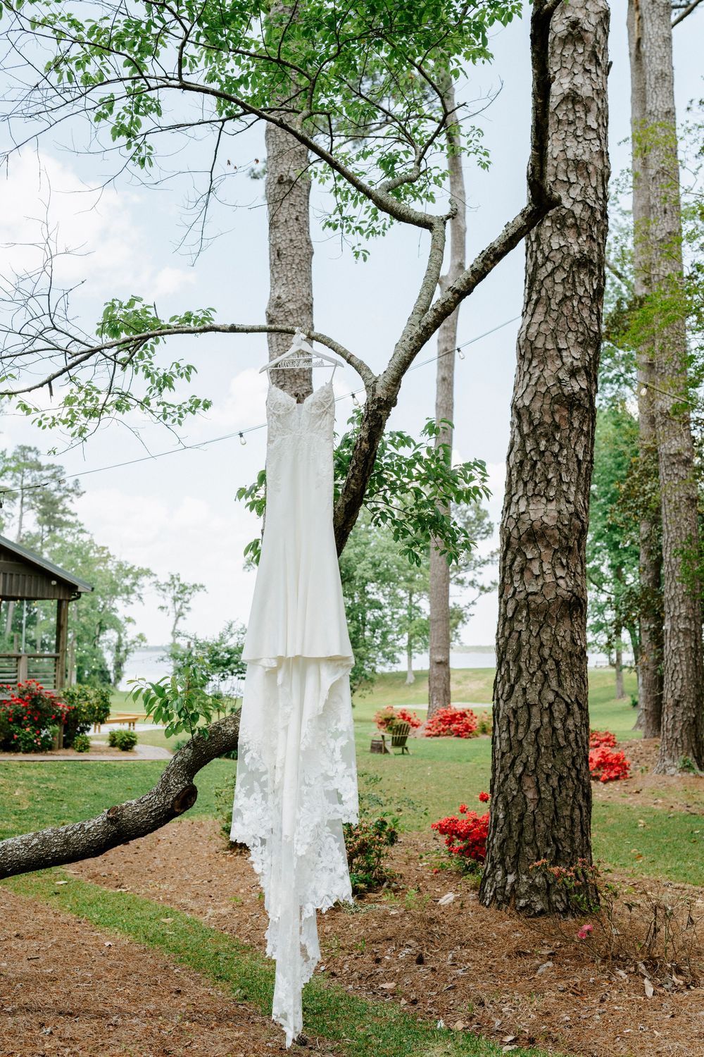 White wedding dress hanging from a tree branch outdoors near a lakeside venue with red flowers.