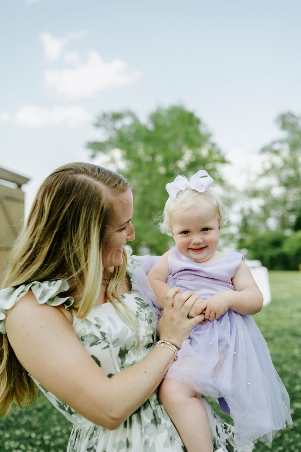 A smiling mother in a floral dress holds her happy baby girl in a purple tutu dress with a white bow outdoors.