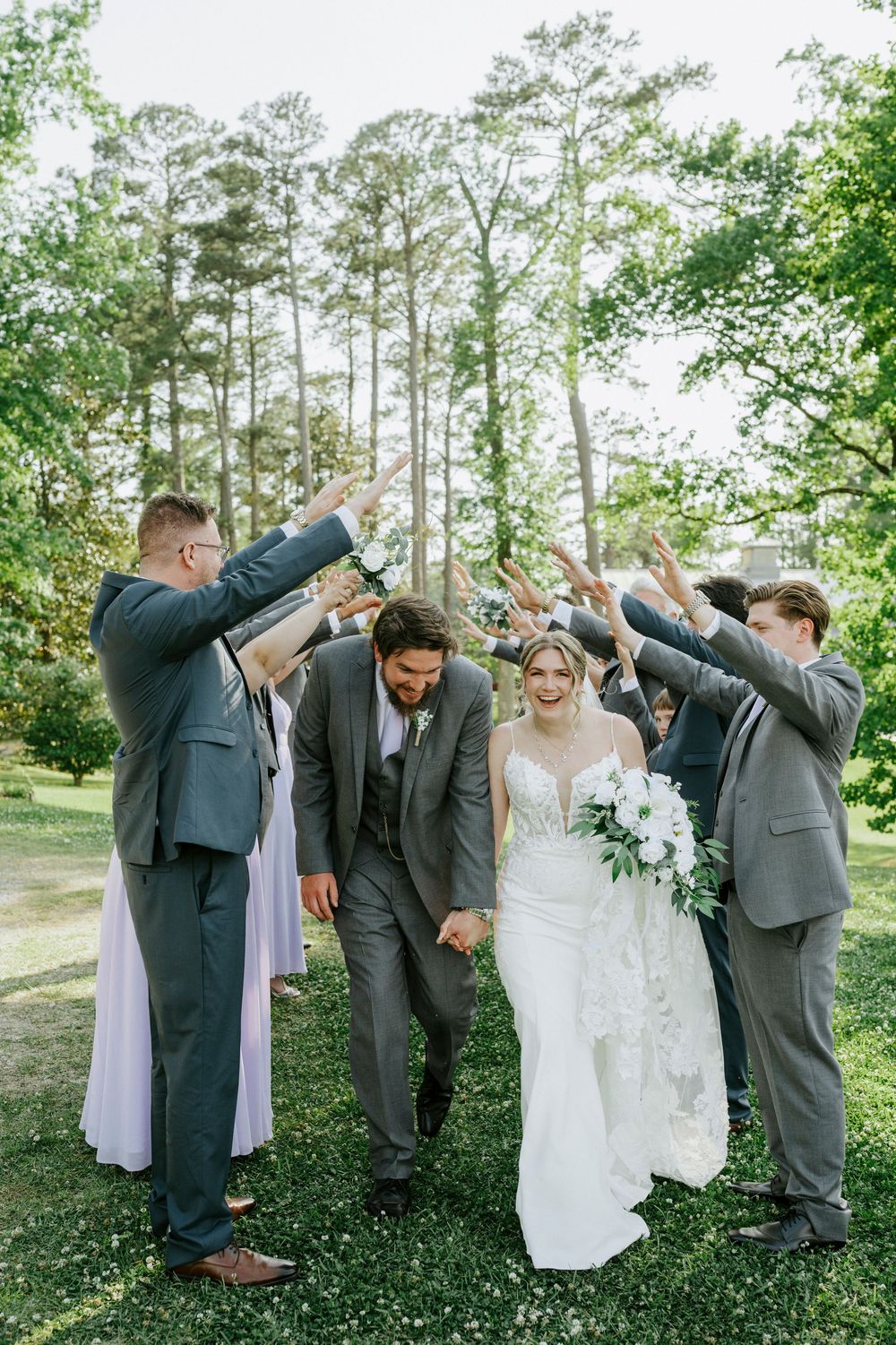 Wedding party celebrates bride and groom walking through arch of raised hands outdoors.