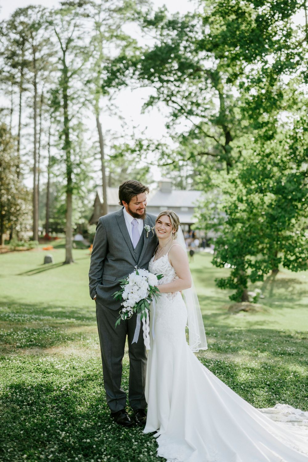 Bride and groom pose together on lush green lawn holding white floral bouquet.