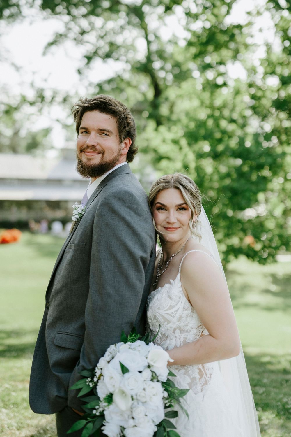 A smiling bride and groom pose back-to-back outdoors, she holds white floral bouquet, wearing lace gown with veil.