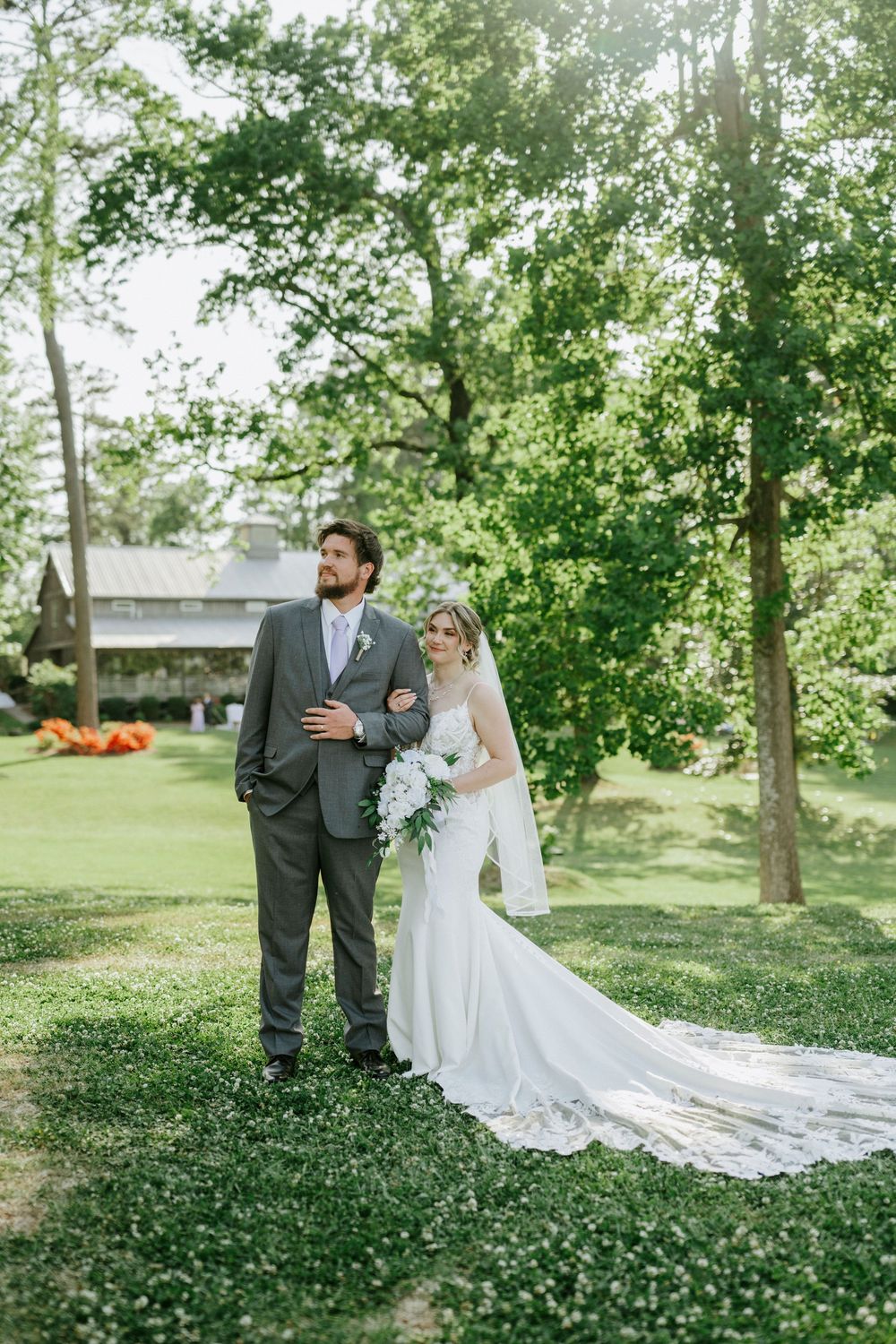 Elegant bride and groom stand on green grass with long cathedral veil trailing.