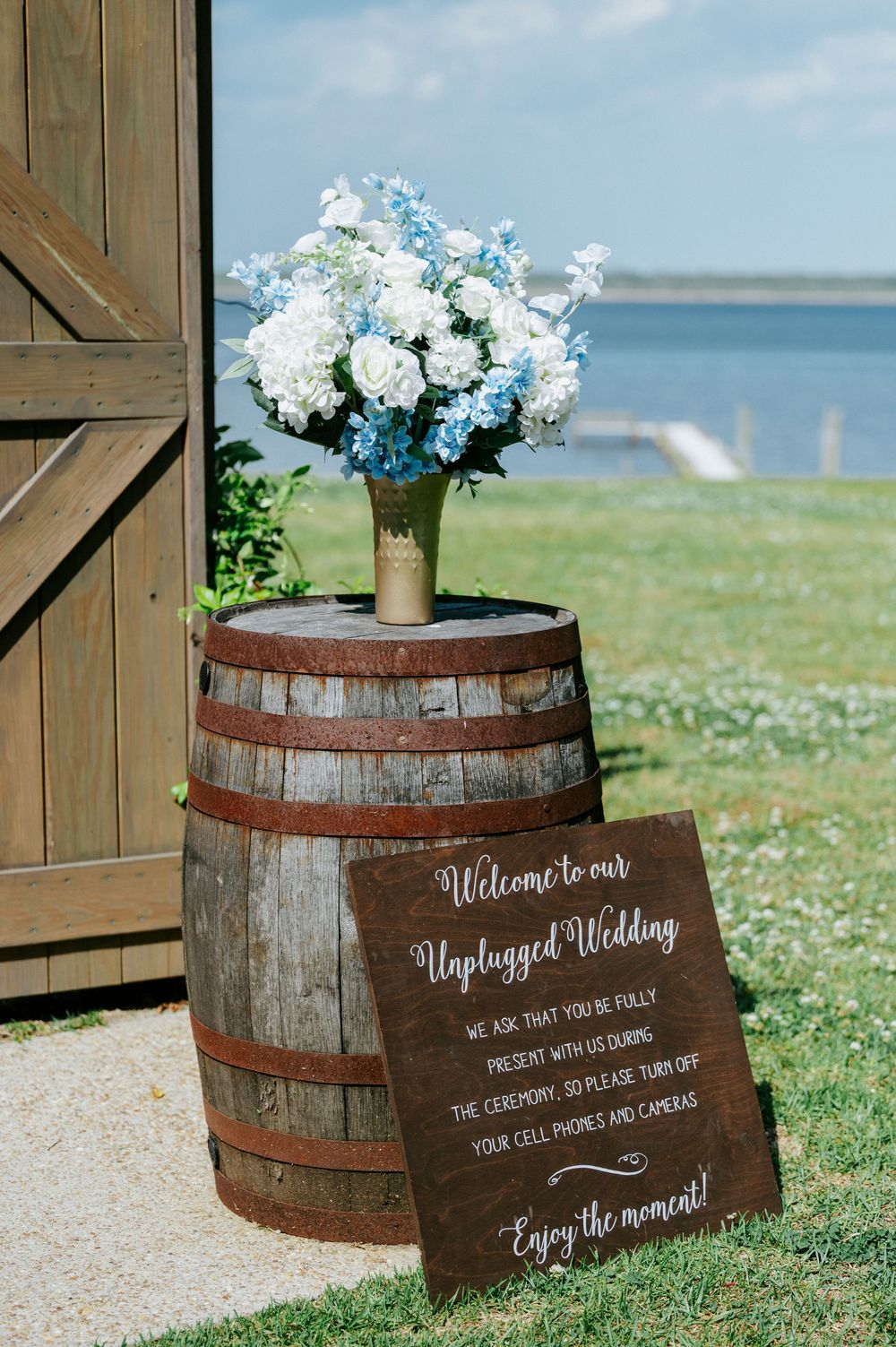 Rustic wedding barrel display with blue and white floral arrangement and unplugged wedding sign near waterfront barn.