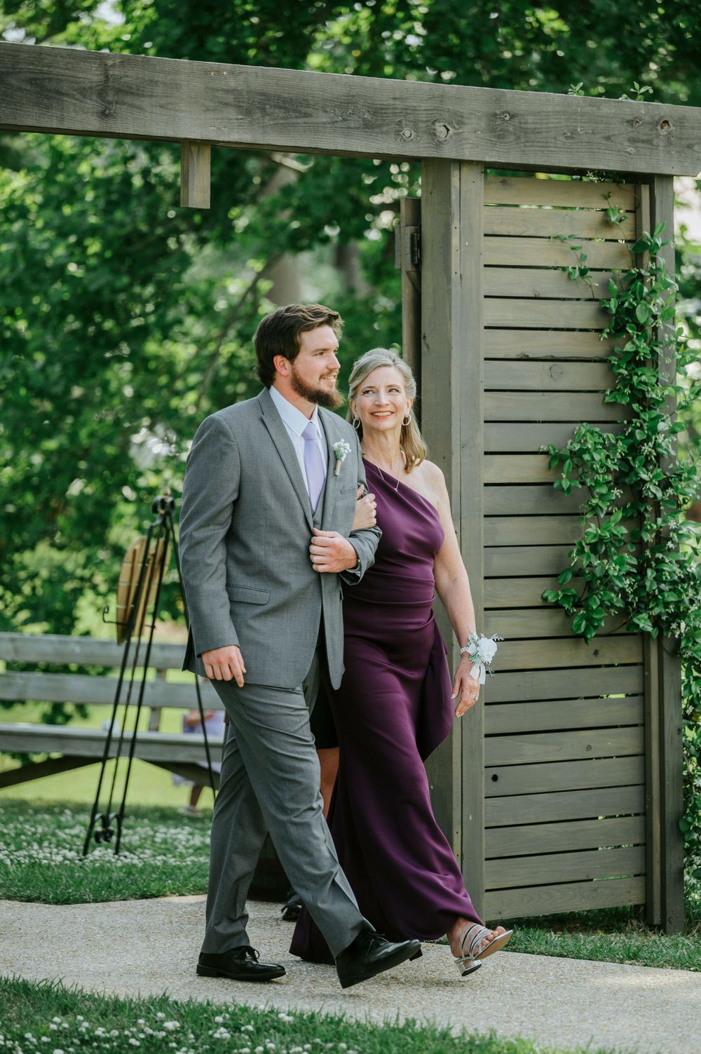 A young man in a gray suit escorts an elegant woman in a purple gown through a garden pergola at a wedding.